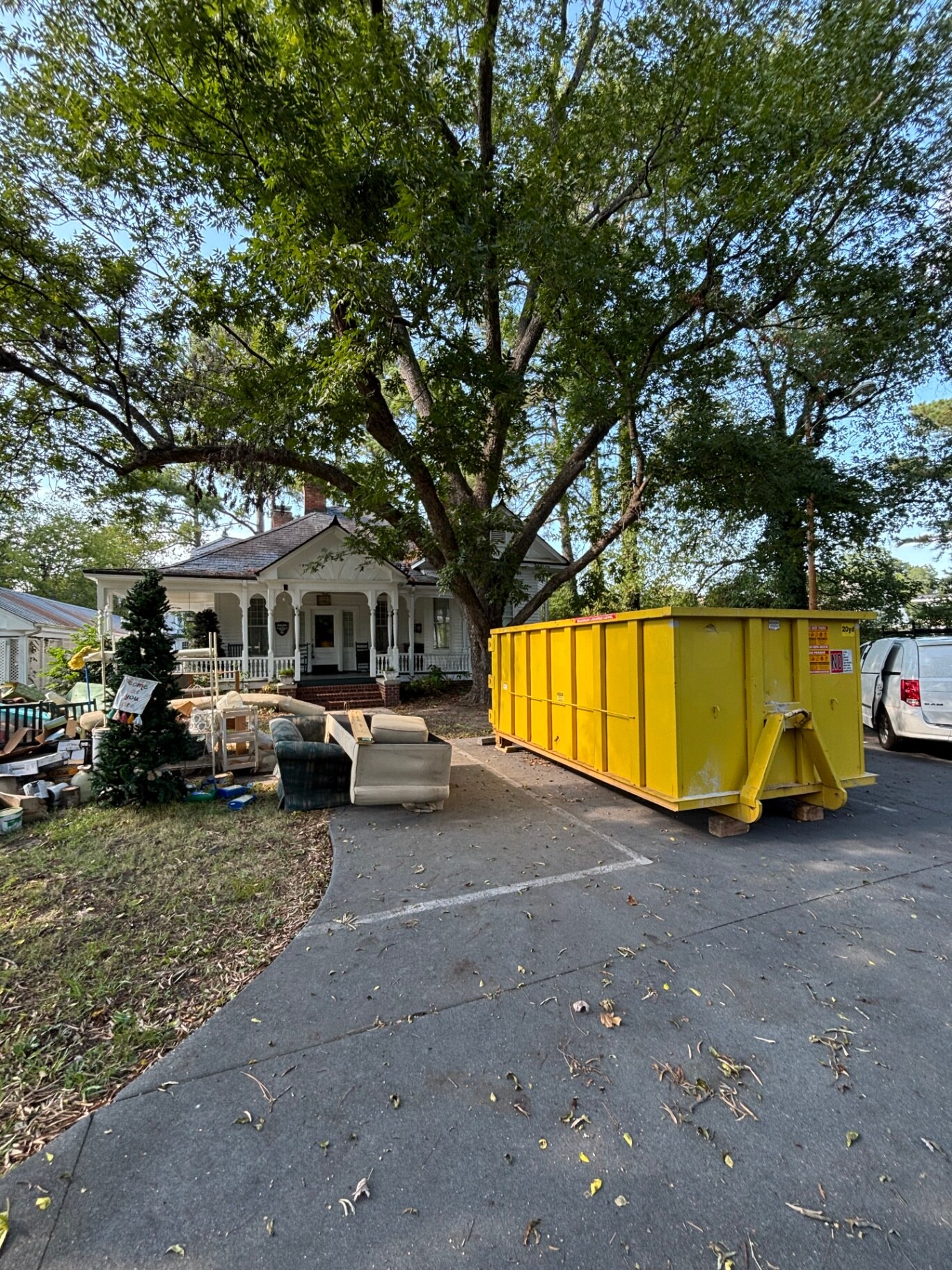 Yellow 20-yard roll-off dumpster on wood blocks in a driveway beside a pile of cleanout junk on the front lawn