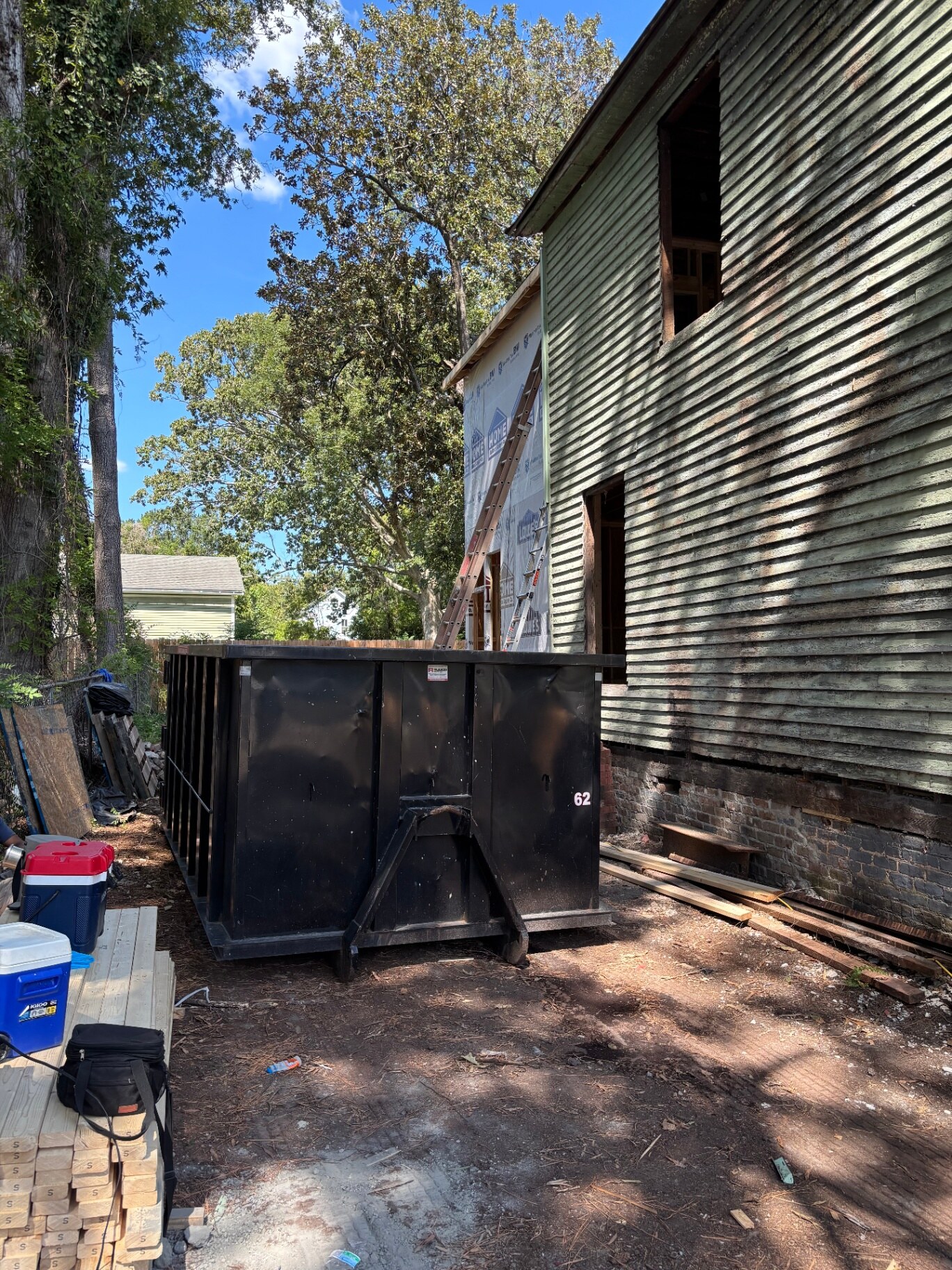 Black roll-off dumpster set beside a two-story wood-sided home under renovation, with ladders and stacked lumber in the yard