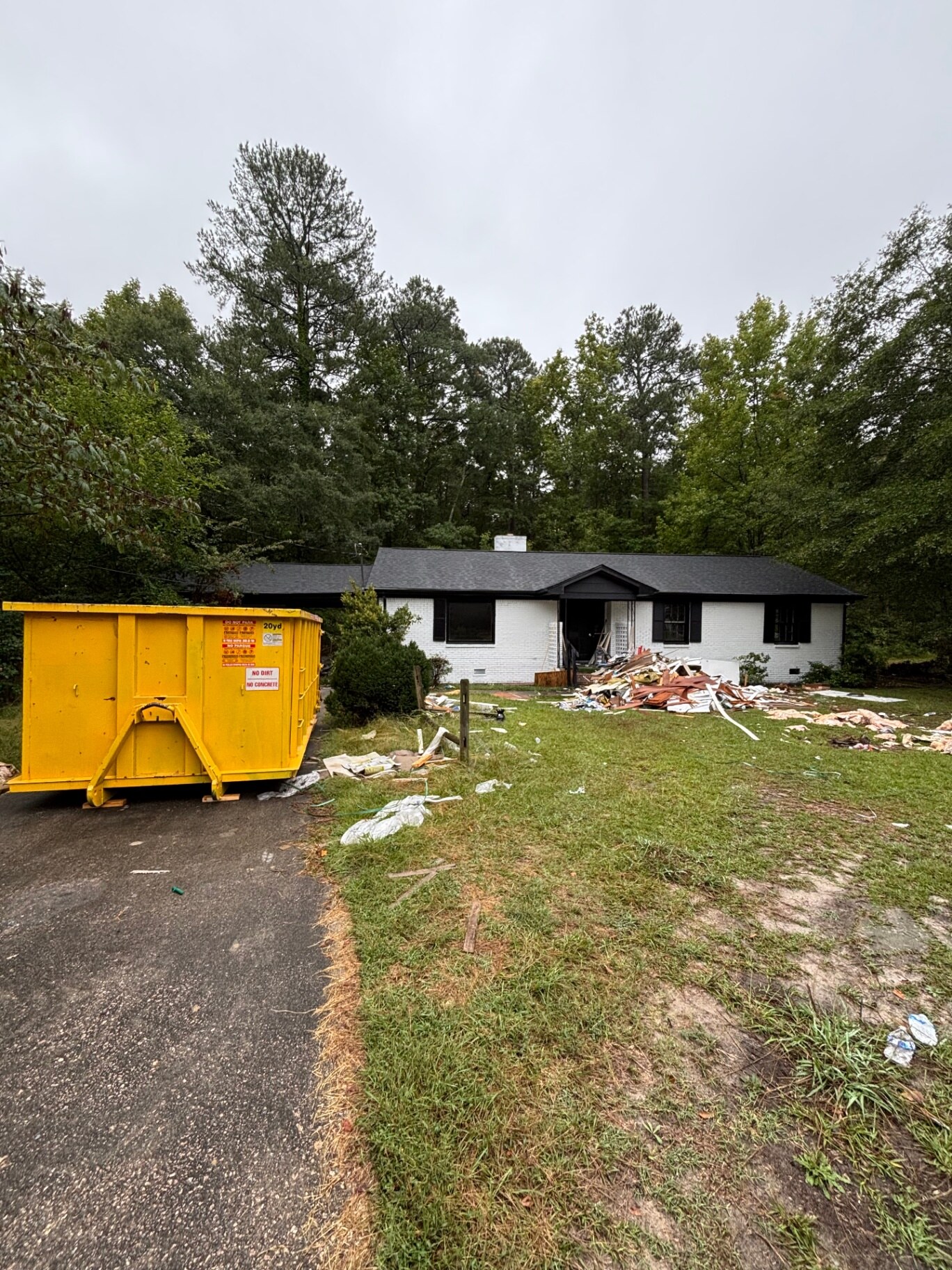 Yellow 20-yard roll-off dumpster on wood blocks in a driveway with a pile of torn-out drywall and framing on the front lawn